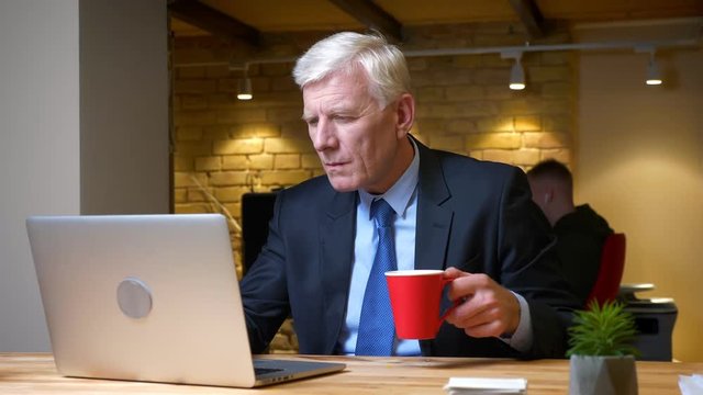 Closeup shoot of old caucasian businessman using the laptop and drinking coffee being busy and focused indoors in the office on the workplace