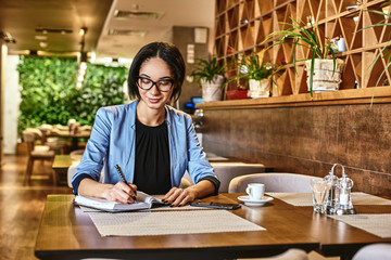 The greatest ability in business is to get along with others and to influence their actions. Young beautiful businesswoman smiling, writing in notebook while working in cafe