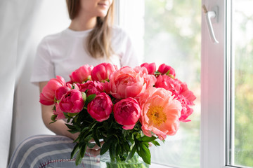 Young girl sitting on the windowsill with Coral peonies in a glass vase. Beautiful peony from...