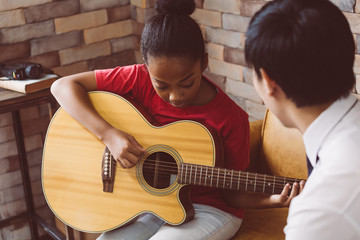 African American girl in casual outfit learning how to play guitar while sitting on couch near male...