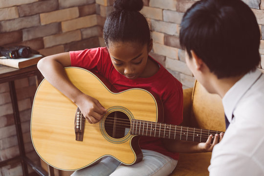 African American girl in casual outfit learning how to play guitar while sitting on couch near male tutor during music lesson