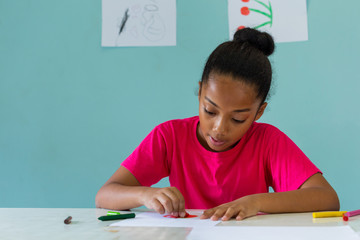 African American girl in bright T-shirt drawing with crayons while sitting at table against blue wall during art lesson at school