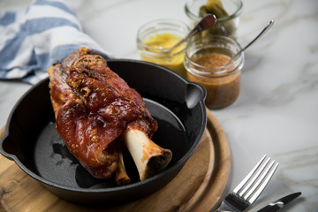 Gegrillte Schweinefleisch Haxe in gusseiserne Pfanne auf Holzbrett mit Cornichons, süßen und mittelscharfen Senf auf Marmor Hintergrund