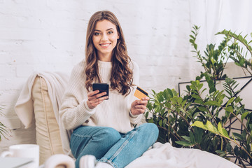 happy pretty girl holding credit card and using smartphone while smiling at camera