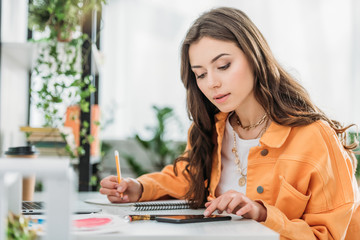 attentive young woman sitting at desk, using smartphone and writing in notebook