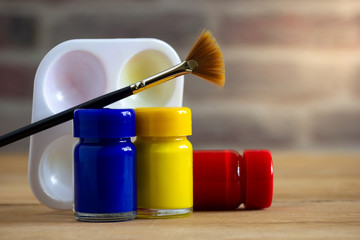 Poster colour bottle, paintbrush and palette on wooden table with brick wall background and morning...