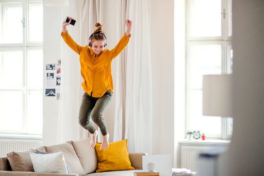 A Young Female Student With Headphones Jumping On Sofa When Studying.