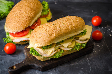 Two delicious sandwiches with ham, cheese, tomatoes, salty cucumber and salad over a wooden board on black background.