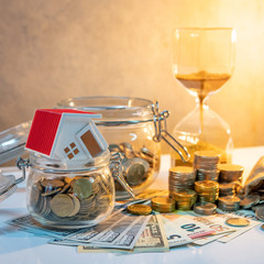 Saving money for retirement concept. Real estate or property investment. Home mortgage loan rate. Coin stack, money bag and currency glass jars on banknotes with hourglasses, house model on the table.