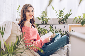 selective focus of cheerful girl holding book and smiling at camera while sitting in room with...