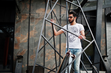 Street style arab man in eyeglasses with longboard posed inside metal pyramid construction.