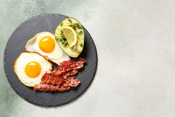 Slate plate with tasty fried eggs, bacon and avocado on light background