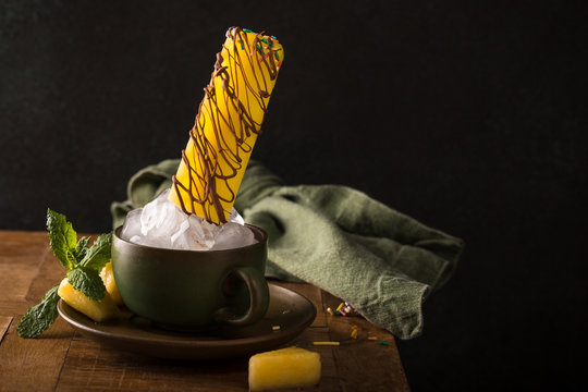 Delicious homemade pineapple in chocolate popsicle in green cup on wooden table. Summer food concept with copy space. Dark photo