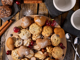 Oatmeal cookies and sand chocolate cake with cherry berry on cutting board on wooden table in rustic style. Top view of still life served with cups and dessert spoons.