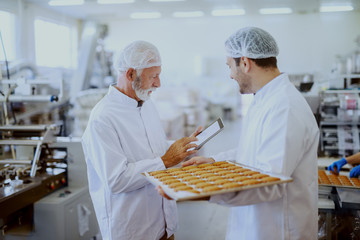 Young employee in sterile white uniform standing with tray with cookies in food plant. Next to him standing supervisor, holding tablet and checking quality of food. - obrazy, fototapety, plakaty