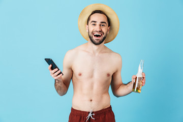 Fototapeta premium Handsome young summer happy man posing isolated over blue wall background drinking beer alcohol using mobile phone.