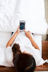 High view image of young woman relaxing in bed and using smart home system on her smartphone to set...