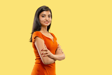 Obraz premium Beautiful female half-length portrait isolated on yellow studio background. Young emotional indian woman in dress standing crossing hands. Negative space. Facial expression, human emotions concept.