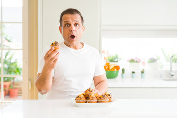 Middle age man eating chocolate chip muffins at home scared in shock with a surprise face, afraid...