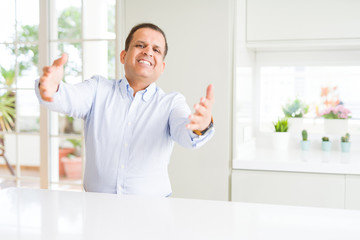Middle age man sitting at home looking at the camera smiling with open arms for hug. Cheerful expression embracing happiness.