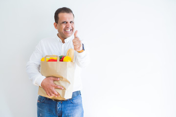 Middle age man holding groceries shopping bag over white background happy with big smile doing ok sign, thumb up with fingers, excellent sign