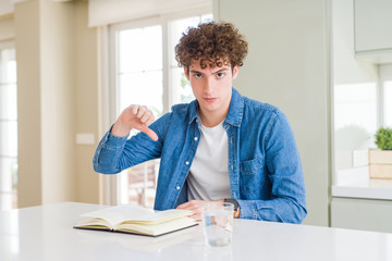 Young man reading a book at home with angry face, negative sign showing dislike with thumbs down, rejection concept