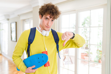 Young student man wearing headphones and backpack holding skateboard with angry face, negative sign showing dislike with thumbs down, rejection concept