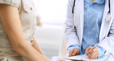 Obraz premium Doctor and patient talking. Physician at work in hospital while writing up medication history records form on clipboard near sitting woman. Healthcare and medicine concepts