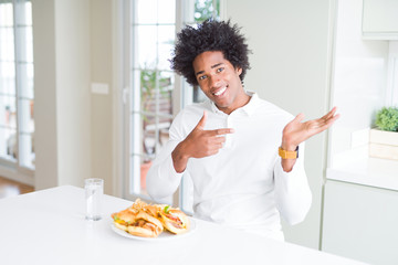Fototapeta na wymiar African American hungry man eating hamburger for lunch amazed and smiling to the camera while presenting with hand and pointing with finger.