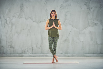 Fototapeta premium Full length of charming fit Caucasian brunette dressed in green outfit standing on mat and doing yoga.