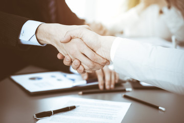 Business people shaking hands at meeting while theirs colleagues clapping and applauding. Group of unknown businessmen and women in modern white office. Success teamwork, partnership and handshake