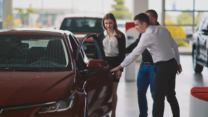Young serious couple talking with handsome shop assistant and trying new car in dealership - Powered by Adobe
