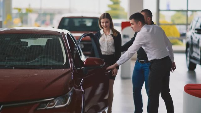 Young serious couple talking with handsome shop assistant and trying new car in dealership