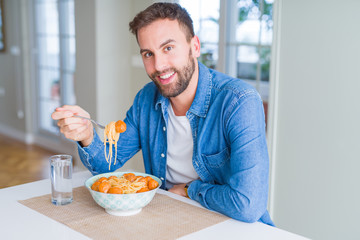 Handsome man eating pasta with meatballs and tomato sauce at home while smiling at the camera