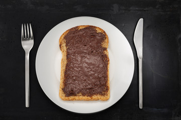 Slice of brown bread with chocolate spread on white plate, stainless steel silverware and black stone background.