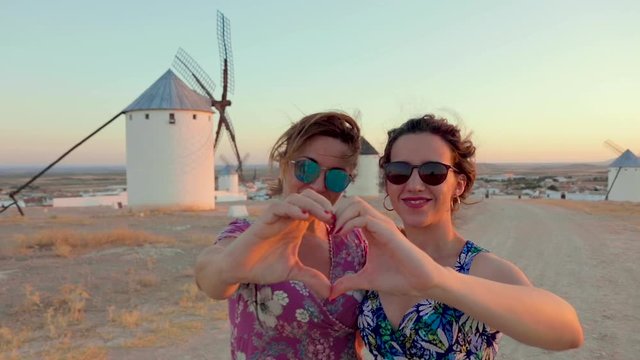 A couple of smiling young women posing for the camera, making a heart with hands, near an old rustic windmill at Castilla La Mancha, Spain