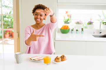 Young african american woman eating breaksfast in the morning at home smiling making frame with hands and fingers with happy face. Creativity and photography concept.
