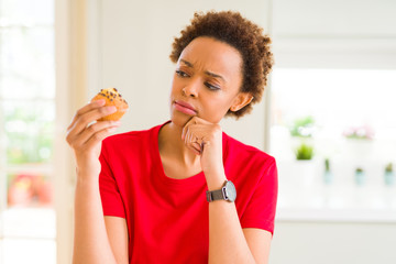 Young african american woman eating chocolate chips muffins serious face thinking about question,...