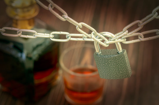 Prohibition of alcohol consumption. The concept of alcoholism. A bottle of cognac and a poured glass in the background. Lock and chains in the foreground. Vignetting photo.