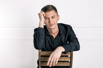 Obraz premium Pensive young handsome man in black shirt sitting on the chair and looking at camera