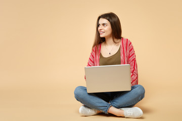 Portrait of young woman in casual clothes looking aside, sitting, using laptop pc computer isolated on pastel beige background in studio. People sincere emotions lifestyle concept. Mock up copy space.