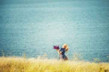 A girl whirling with a bouquet of wild flowers in her hands among the grass on a cliff by the sea. The atmosphere of happiness and carelessness.