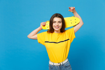 Portrait of smiling young woman in vivid casual clothes standing, looking camera, holding yellow skateboard isolated on blue wall background in studio. People lifestyle concept. Mock up copy space.