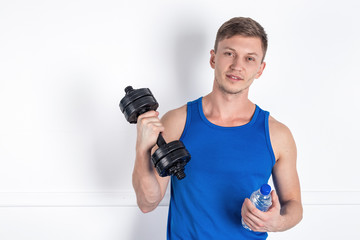 Fototapeta na wymiar Handsome young man in blue shirt holding a dumbbell and a water bottle. White background