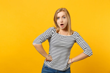 Excited young woman in striped clothes keeping mouth open standing with arms akimbo isolated on yellow orange wall background in studio. People sincere emotions, lifestyle concept. Mock up copy space.