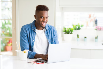 Handsome young african business man eating delivery asian food and working using computer, enjoying noodles smiling