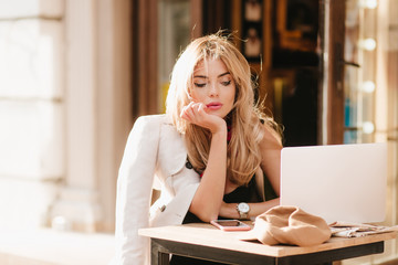 Frustrated fair-haired woman waiting for call while working with computer in outdoor cafe. Tired business-lady wrapped in coat using laptop and thinking about something unpleasant.
