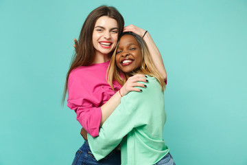 Two young women friends european and african american in pink green clothes standing posing isolated on blue turquoise wall background, studio portrait. People lifestyle concept. Mock up copy space.