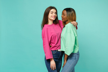 Two young women friends european and african american in pink green clothes standing posing isolated on blue turquoise wall background, studio portrait. People lifestyle concept. Mock up copy space.