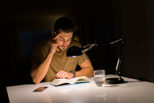 Young handsome man studying at home, reading a book at night